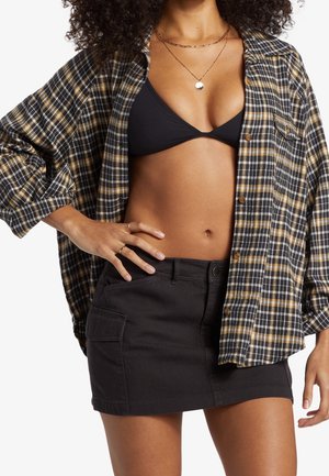 Donna che indossa una camicia a quadri aperta sopra un top bikini nero e una minigonna nera, con la mano appoggiata sull'anca e collane a strati.