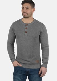 Maglione grigio a maglia con texture a coste, scollo rotondo, tre bottoni marroni decorativi e un'etichetta marrone sul lato inferiore.