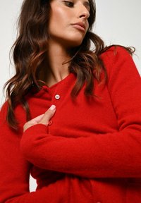 Czerwony sweter z dzianiny z dekoltem w kształcie litery V, długimi rękawami i zapięciem na guziki. Miękka faktura oraz luźny krój, prezentujący prosty design.