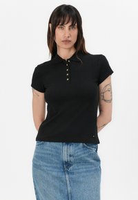 Zwarte polo met een kraag, korte mouwen en gouden knoopaccenten. Gemaakt van een zachte stof, gecombineerd met een dennenrok.