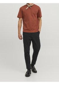 T-shirt rosso ruggine a maniche corte con colletto rotondo e un piccolo logo stampato in nero, abbinato a pantaloni neri a sigaretta e mocassini neri.