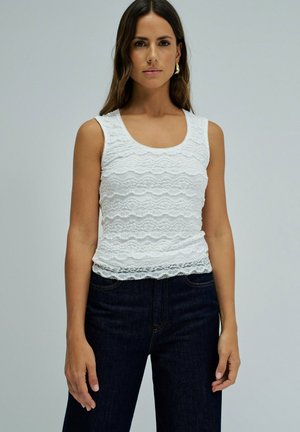 Femme aux longs cheveux portant un haut sans manches en dentelle blanche et un jean bleu foncé, debout devant un fond uni.