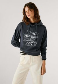Felpa corta blu scuro con grafica testuale e accenti di stelle, dotata di cappuccio con cordoncino e polsini a coste. Indossata con pantaloni chiari.