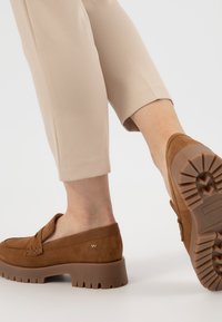 Mocassini in suede marrone con suola in gomma spessa, caratterizzati da un accento cucito e un dettaglio con il logo, abbinati a pantaloni beige a vita corta.
