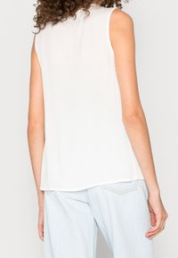 Haut sans manches blanc avec une texture lisse, col rond et un ourlet légèrement incurvé, associé à un jean bleu clair.