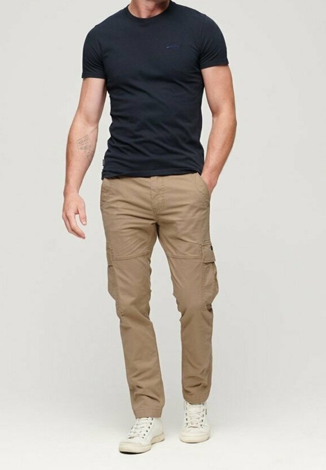 Homme portant un t-shirt noir ajusté à manches courtes, un pantalon cargo beige avec des poches latérales, et des baskets blanches, debout avec une main dans la poche.