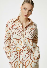 Button-up shirt in light beige, featuring a bold rose pattern in rust and golden hues. Flap collar and long sleeves with button cuffs.