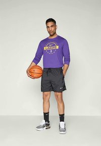 Paarse lange mouwen shirt met "LAKERS" logo, zwarte shorts en zwart-witte sneakers. Houdt een basketbal vast, terwijl hij op een neutrale achtergrond staat.