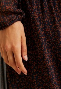 Main gauche avec du vernis à ongles rose clair reposant contre un tissu foncé à motif abstrait de feuilles marron sur une manche froncée.