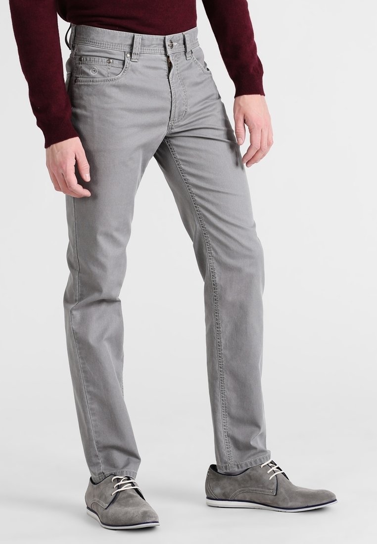 Homme portant un jean slim gris clair, des chaussures en daim grises avec des lacets blancs, et une chemise à manches longues bordeaux, debout devant un fond blanc.