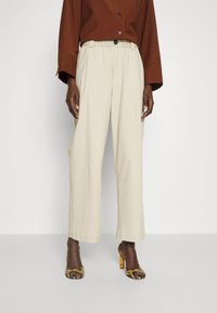 Beżowe, szerokie spodnie z elastycznym pasem i zapięciem na guzik, w połączeniu z brązową bluzką i wzorzystymi butami na obcasie.