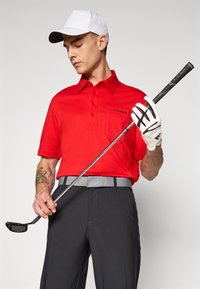 Cămașă polo roșie cu guler, mâneci scurte și un buzunar pe piept, asortată cu pantaloni gri și o curea gri. Ținând un băț de golf, purtând o șapcă albă și mănuși.