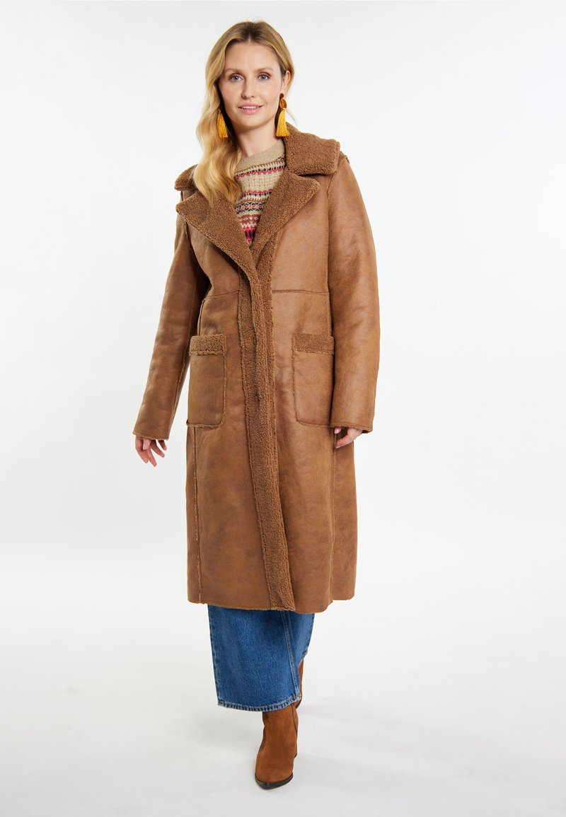 Brun, lang frakke med en shearling-krave, to frontlommer og en struktureret overflade. Båret over en mønstret sweater og en blå denimnederdel.