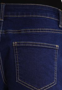 Donkerblauwe spijkerbroeken met de nadruk op de achterzak en broeklussen, met zichtbare beige stiksels langs de naden.