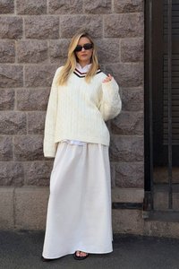 Cremfarbener Kabelstrickpullover mit einem schwarz-roten V-Ausschnitt, über einer weißen Bluse getragen und kombiniert mit einem fließenden weißen Maxirock und flachen Sandalen.