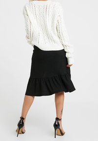 Vit stickad tröja med ett kabelm�önster, tillsammans med en svart knä-lång kjol med volangkant och högklackade skor.