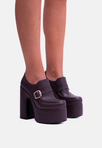 Scarpe con zeppa marroni e tacco spesso, realizzate in pelle liscia. Caratterizzate da una cintura con fibbia e una punta arrotondata.