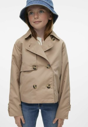 Beige dobbeltradet frakke med store knapper og spids krave, båret over en hvid top og parret med blå jeans. Blå denim bøttehat.