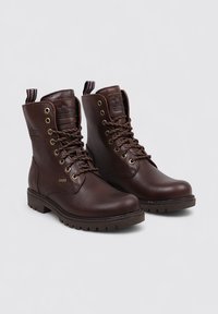 Botas de cuero marrón hasta el tobillo con punta redonda, ojales de metal, cordones marrones y una superficie texturizada. Presenta una etiqueta con el logo en el talón y una suela robusta.