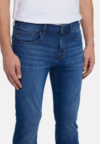 Jeans in denim blu con un design aderente, cinque tasche, dettagli di cucitura discreti e un taglio dritto. Indossati con una t-shirt bianca.