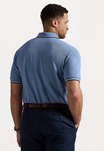 Πόλο μπλουζάκι σε απαλό μπλε με υφή, κοντά μανίκια και αντίθετη σκοτεινή διακόσμηση γύρω από τον γιακά. Φορεμένο με σκούρο παντελόνι και καφέ ζώνη.