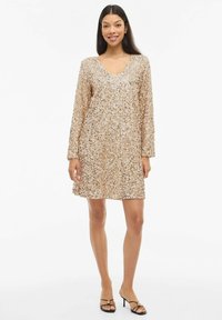 Beżowa sukienka z cekinami, z długimi rękawami, dekoltem w szpic i sylwetką w kształcie litery A. Charakteryzuje się całościowo połyskującą teksturą i długością do kolan.