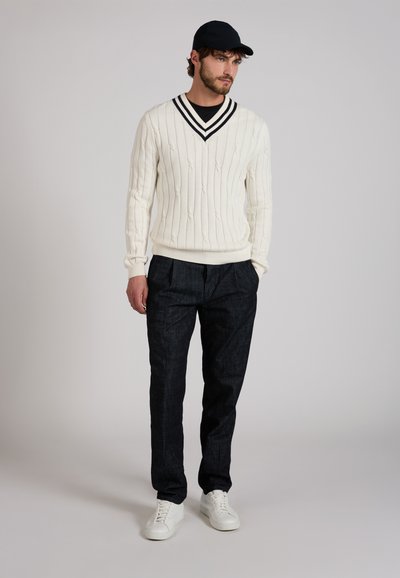 Man wearing white V-neck cable knit sweater, black cap, dark pants, and white sneakers, standing with hands in pockets on gray background.