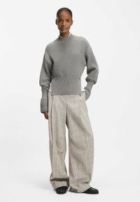 Woman wearing a gray cropped knit sweater, wide-leg beige plaid trousers, black shoes, standing with hands in pockets against white background.