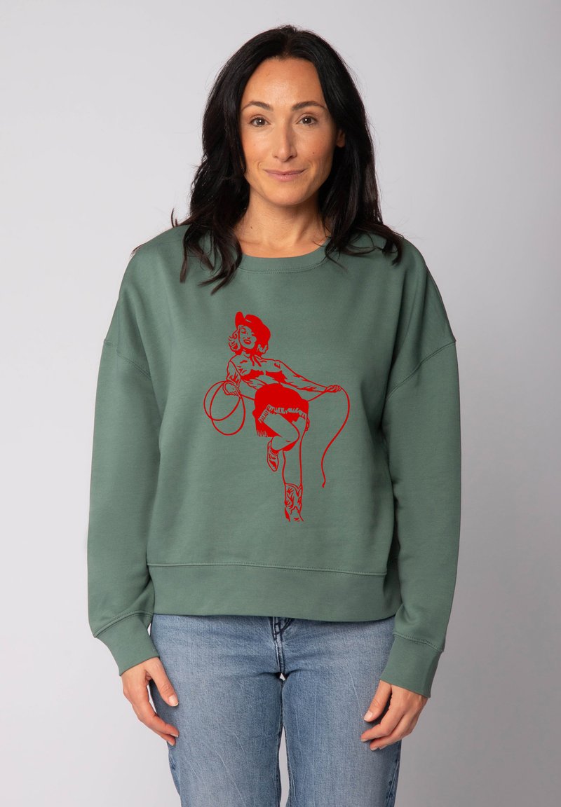 Grüner Pullover mit einem roten Grafikdruck einer Frau, die einen Lasso hält. Hat eine lockere Passform, Rundhalsausschnitt und Rippbündchen.