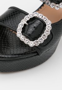 Sandales à plateforme noires en cuir texturé, dotées d'une boucle ronde ornée de strass et d'accents lisses. La semelle présente une finition à motif.