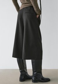 Culottes noires en tissu texturé, dotées d'une taille haute, de poches latérales et d'une coupe ample, associées à des bottes noires montantes jusqu'au genou.