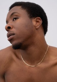 Halskette aus goldfarbenem Metall mit einem flachen, länglichen Design. Die Kette hat eine glänzende Oberfläche und wird auf der nackten Haut getragen.