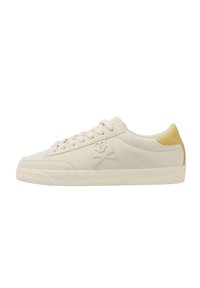 Sneaker in camoscio beige con suola in gomma bianca, caratterizzata da un accento giallo sul tallone e un logo di teschio e ossa incrociate sul lato.