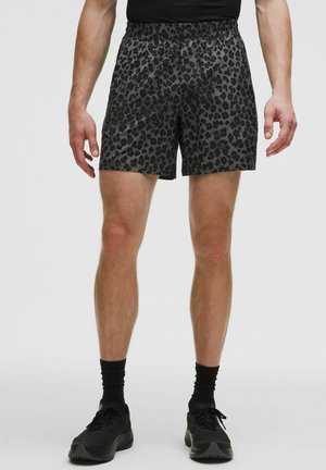 Mann trägt schwarze und dunkelgraue Sportshorts mit Leopardenmuster, schwarze Socken und schwarze Laufschuhe, steht vor einem einfarbigen Hintergrund.