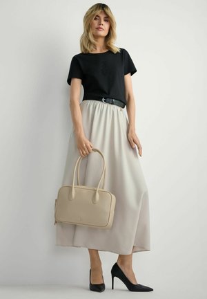 Kobieta ubrana w czarną bluzkę z ozdobami, beżową długą spódnicę, czarne szpilki i trzymająca beżową torebkę na białym tle.