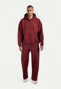 Vinröd zip-up hoodie med en framficka och matchande löst sittande byxor. Mjukt tyg, ribbade muddar och vita sneakers kompletterar looken.