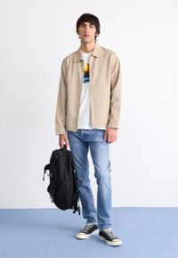 Giacca leggera beige, t-shirt bianca con grafica colorata, jeans blu, zaino nero e sneakers nere in tela. Design semplice, vestibilità casual.