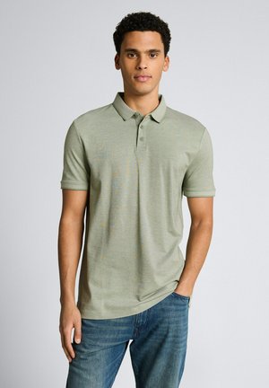 Uomo con capelli ricci che indossa una polo verde chiaro a maniche corte e jeans blu, in piedi con una mano in tasca davanti a uno sfondo semplice.