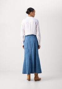 Denim maxi rok met een uitlopende vorm, blauwe kleur en voornaad. Gecombineerd met een wit linnen overhemd met lange mouwen en een paar bruine enkellaarsjes.