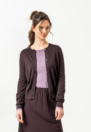 Junge Frau mit welligem Haar zu einem hohen Pferdeschwanz gebunden, trägt ein lila Top und eine dunkelviolette, geknöpfte Strickjacke sowie einen Rock, steht vor weißem Hintergrund.