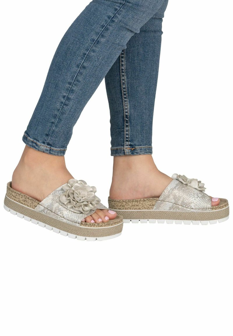 Füße mit hellbeigen Slide-Sandalen mit floralem Dekor oben und weißen Sohlen, kombiniert mit hochgekrempelten blauen Jeans.