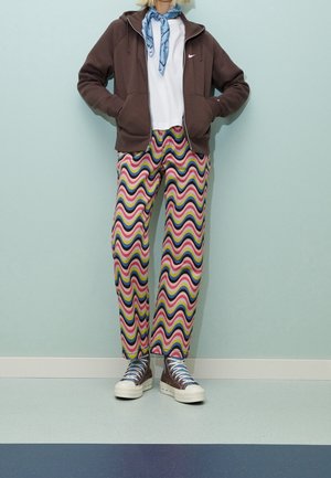 Person wearing a brown zip-up hoodie, white t-shirt, blue scarf, multicolored wavy-patterned pants, and brown high-top sneakers.