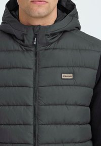 Tumehall padi vest, millel on kapuuts, ja millel on tikitud tekstuur ning lukuga eesmine sulgemine. Rinnal on nähtav väike "Blend" logo.