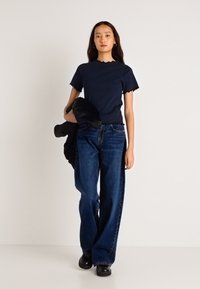 Αυτοκόλλητο ναυτικό μπλουζάκι με κομμένη άκρη συνδυασμένο με ψηλόμεσα, φαρδιά σκούρα μπλε τζιν και μαύρα παπούτσια χωρίς κορδόνια, στέκεται απέναντι σε ουδέτερο φόντο.