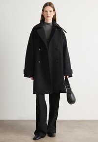 Cappotto nero doppiopetto con ampio colletto, grandi bottoni e vestibilità rilassata. Indossato sopra un dolcevita grigio e abbinato a pantaloni neri.
