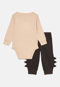 Beige langärmliger Babybody mit schwarzen Hosen, die an den Seiten gestrickte Dinosaurierstachel haben.