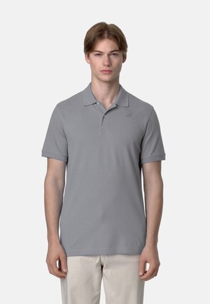 Giovane uomo con capelli castano chiaro che indossa una polo grigia a maniche corte e pantaloni beige chiaro, in piedi davanti a uno sfondo semplice.