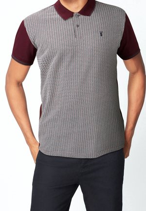 Homme portant un polo à manches courtes avec col et manches bordeaux, panneau avant à petits carreaux, associé à un pantalon noir.
