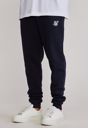 Person trägt marineblaue Jogginghose mit weißem Logo am Oberschenkel und weiße Turnschuhe, steht auf hellgrauem Boden vor hellgrauem Hintergrund.