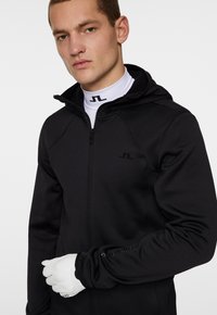 Svart zip-up jacka med huva och texturerat tyg, med en liten logotyp på bröstet och vita handskar, som framhäver en strömlinjeformad design och praktisk funktion.